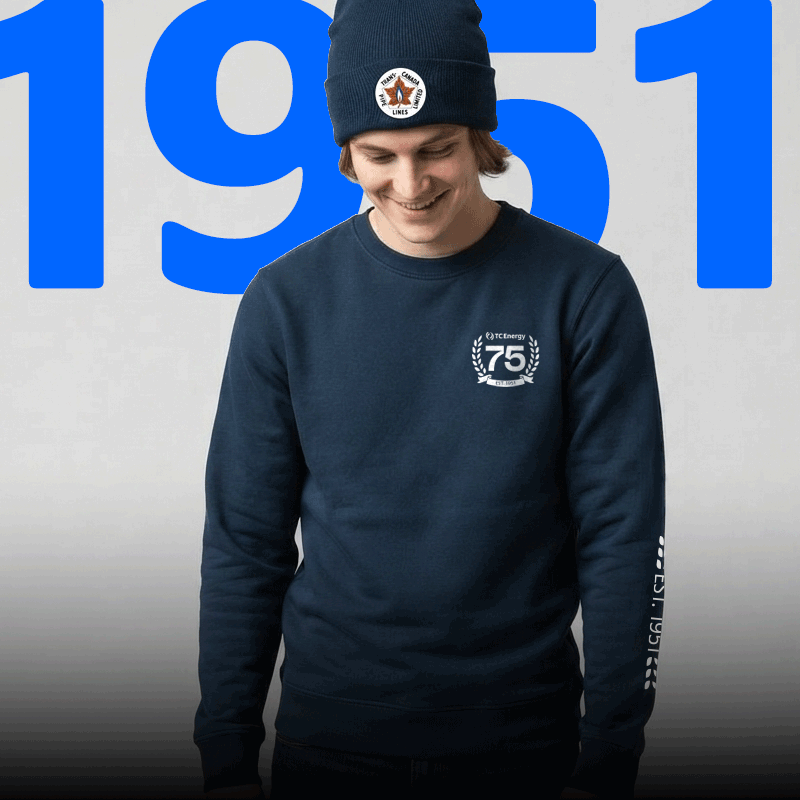 Person wearing a navy beanie and navy crewneck sweatshirt featuring the Trans Canada PipeLines 1951 anniversary logo, standing in front of a large blue 1951 graphic that counts up to 2026