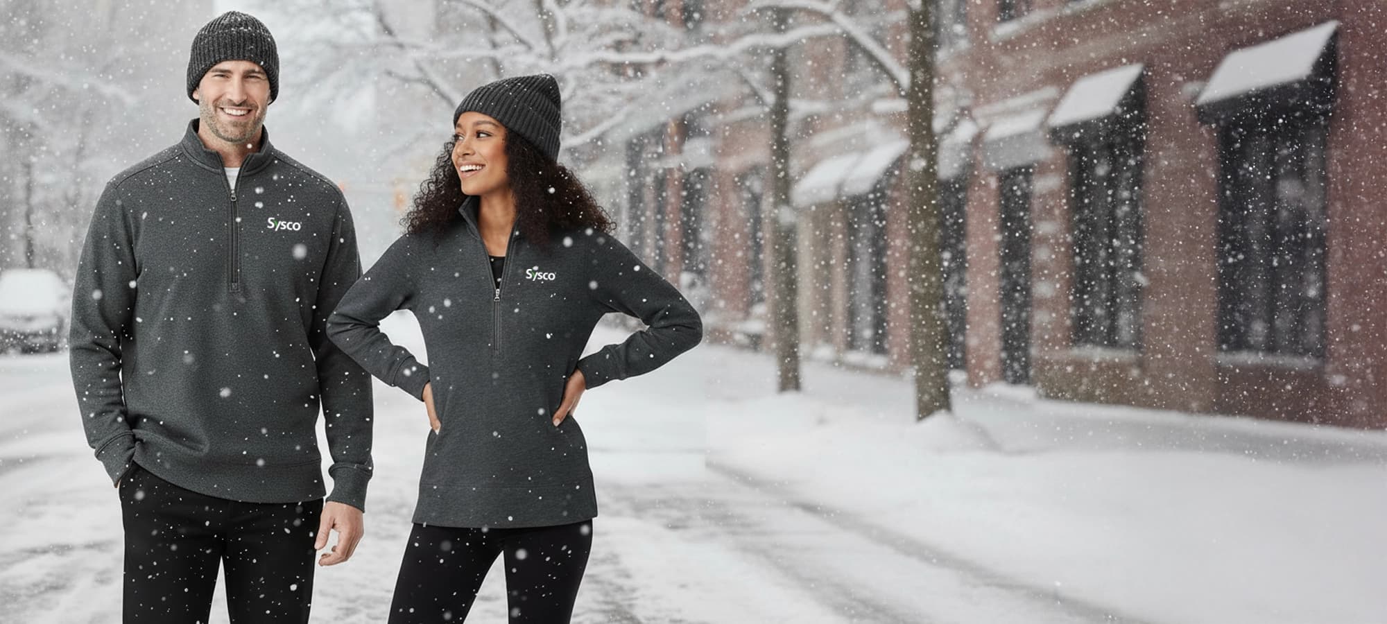 A man and a woman stand outside in the snow, both wearing matching gray quarter-zip pullovers with the Sysco logo, black pants, and gray knit beanies. The man is on the left, smiling at the camera, and the woman is on the right, looking towards the man and smiling. Snowflakes are visible in the foreground, and a blurred street scene with lights is in the background.
