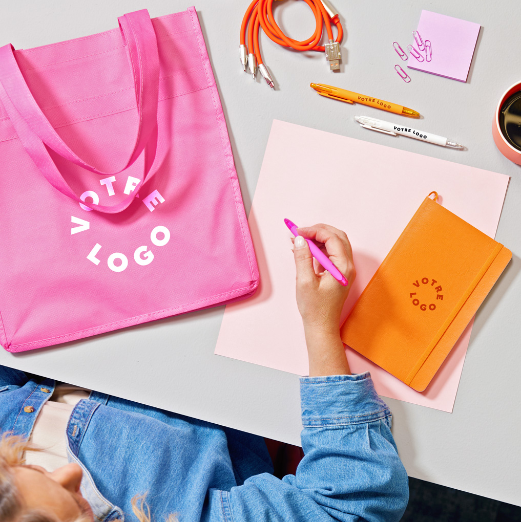 Flat lay of a branded pink tote, pens, notebook, chargers, and other office promo items