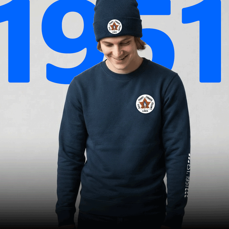 Person wearing a navy beanie and navy crewneck sweatshirt featuring the Trans Canada PipeLines 1951 anniversary logo, standing in front of a large blue 1951 graphic that counts up to 2026