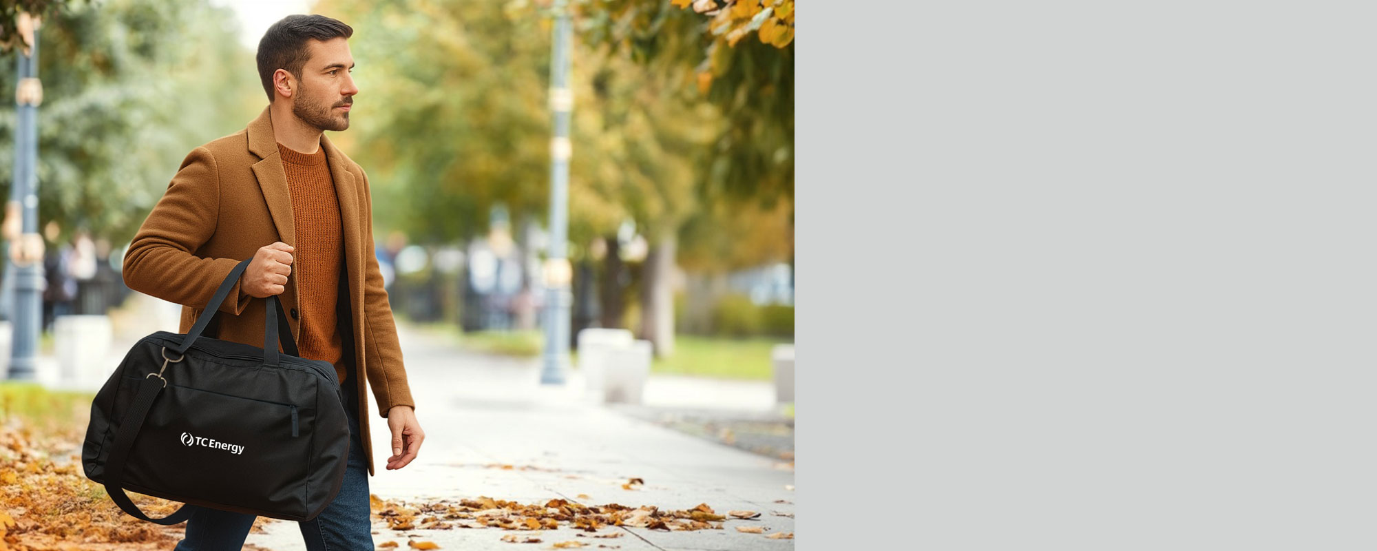 Person walking outdoors on a sidewalk lined with autumn leaves, wearing a brown coat and carrying a black duffel bag with the logo TC Energy
