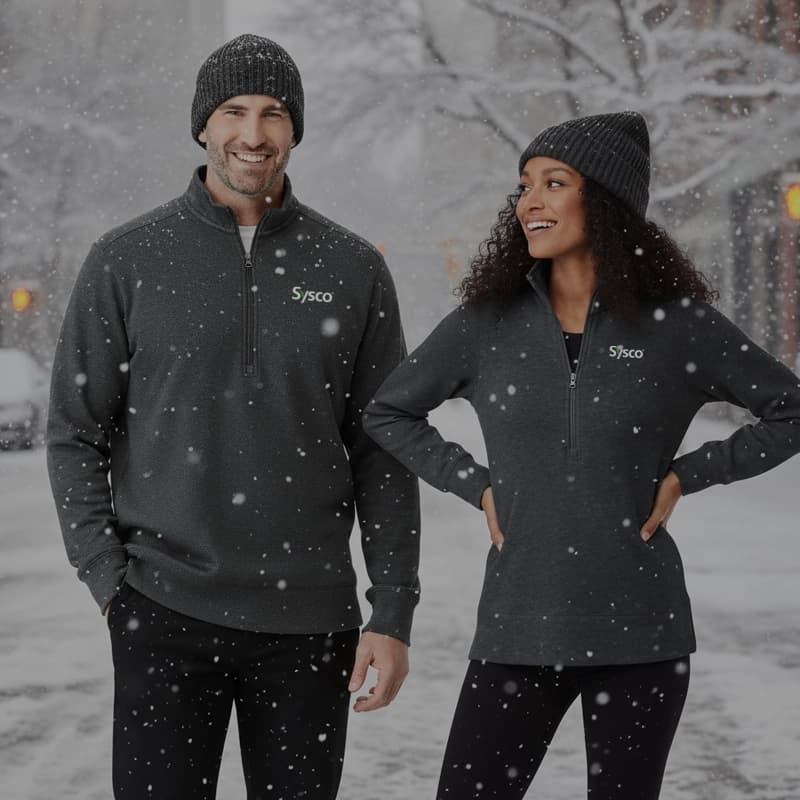 A man and a woman stand outside in the snow, both wearing matching gray quarter-zip pullovers with the Sysco logo, black pants, and gray knit beanies. The man is on the left, smiling at the camera, and the woman is on the right, looking towards the man and smiling. Snowflakes are visible in the foreground, and a blurred street scene with lights is in the background.