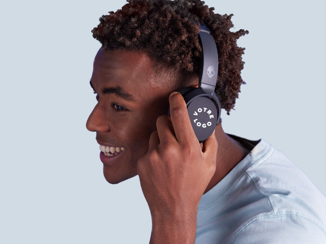 Smiling man with curls wears black Skullcandy headphones printed with YOUR LOGO, represents custom tech gear.