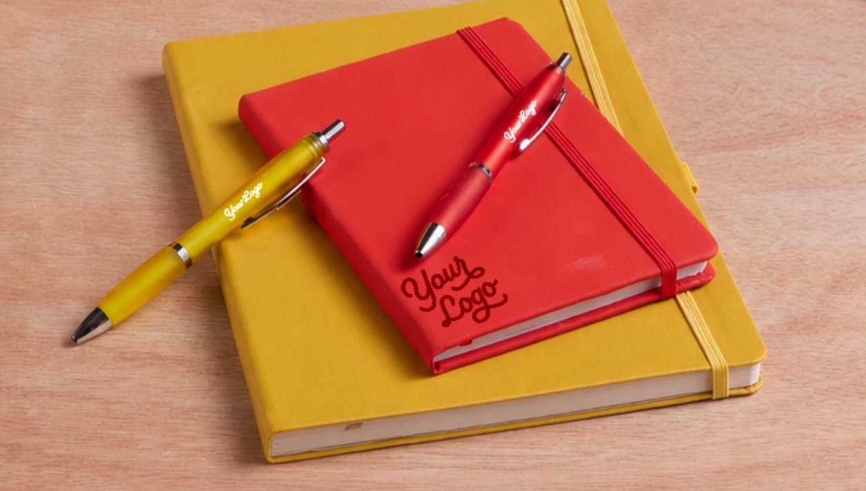 A red journalbook stacked on top of a yellow journalbook, each with a matching pen - all products are branded with Your Logo to show personalization