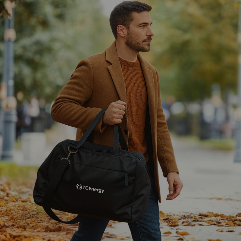 Person walking outdoors on a sidewalk lined with autumn leaves, wearing a brown coat and carrying a black duffel bag with the logo TC Energy