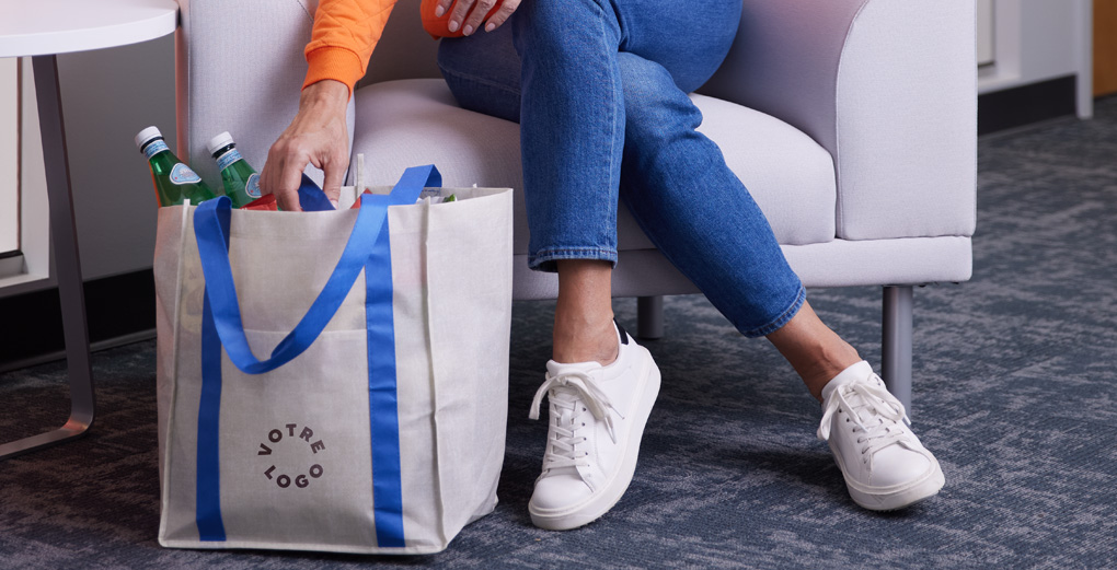 Person in white sneakers places drinks into a canvas tote with blue handles and YOUR LOGO — labeled Tote Bags.