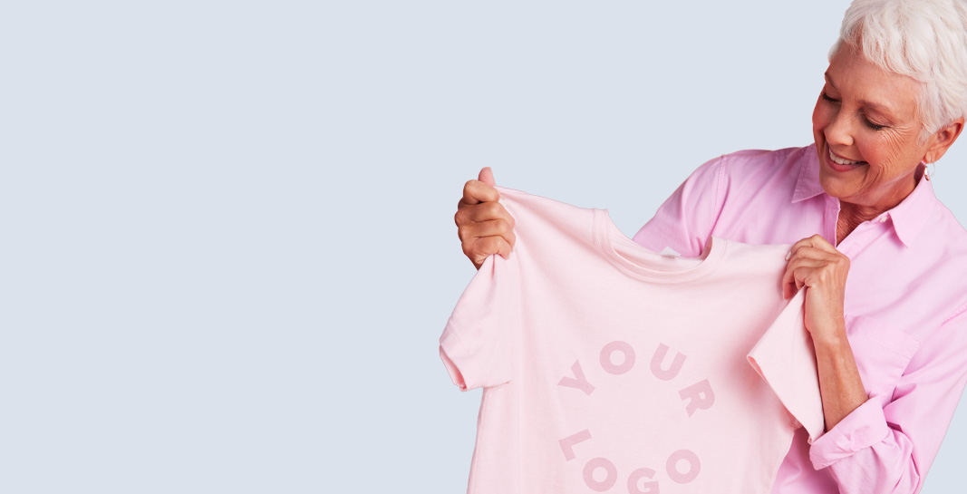Older woman in pink holds YOUR LOGO tee with ORDER ONE... OR MORE! speech bubble and Deals button — for promo deals