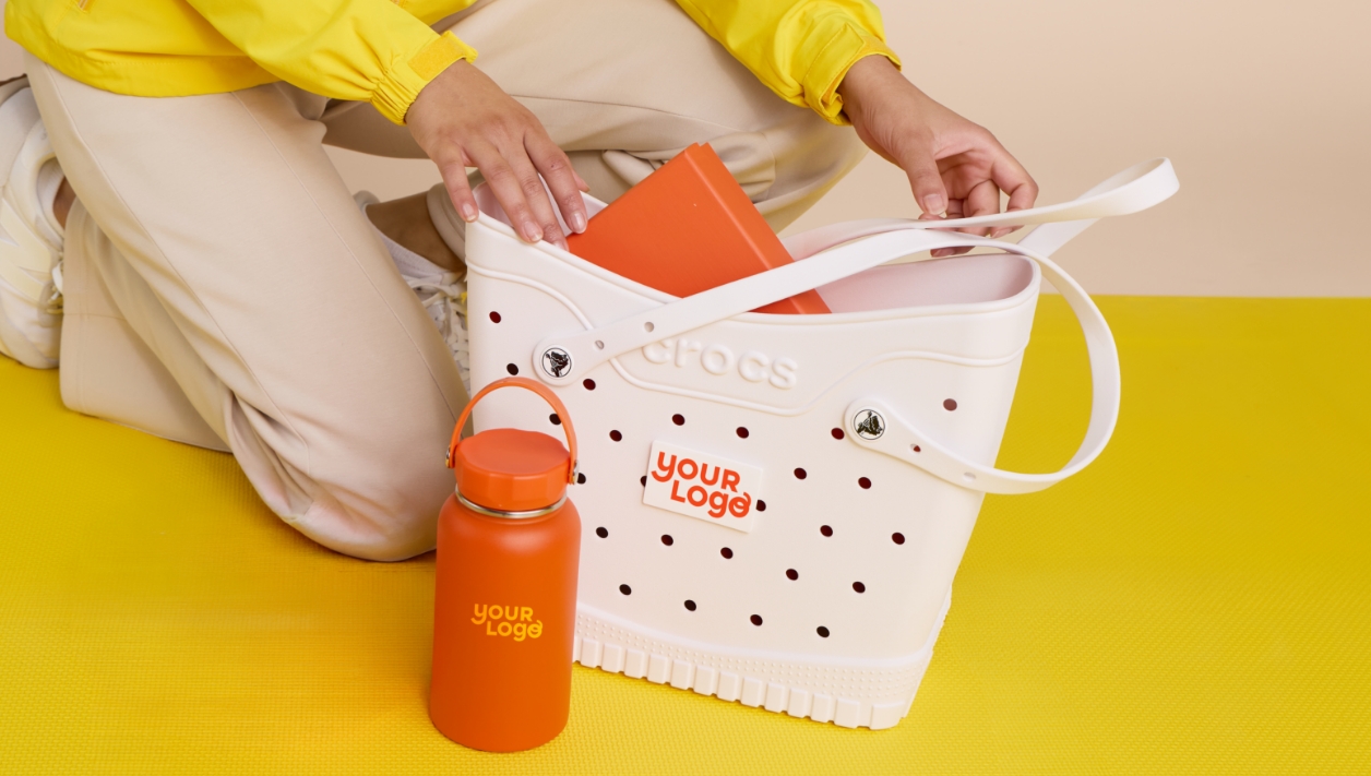 A white Crocs tote bag personalized with Your Logo - a woman is reaching into the bag to pull out other branded merchandise